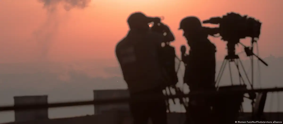 Periodistas palestinos trabajan con una imagen de un ataque aéreo israelí de fondo. Foto:D W.