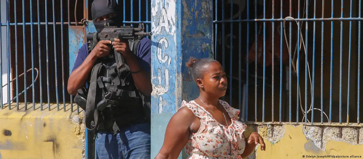 Mujer pasa frente a la vacía Prisión Nacional en Puerto Príncipe, Haití mientras un agente apunta con una metralleta sobre la cabeza de la transeúnteImagen: Odelyn Joseph/AP/